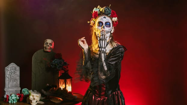 Goddess of death blowing air kisses in studio, acting flirty and romantic with cavalera catrina halloween costume. Doing kissy face with skull body art to celebrate day of teh dead holiday.