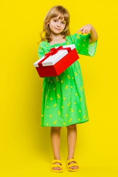Lovely smiling young preteen child girl kid presenting birthday gift box stretches out hands, offer wrapped present career bonus, celebrating party. Little cute children on yellow background. Vertical