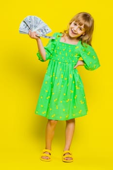 Rich pleased young cute school girl waving money dollar cash banknotes bills like a fan, success, lottery winner, big income, wealth. Teen blonde female child kid on studio yellow background. Vertical