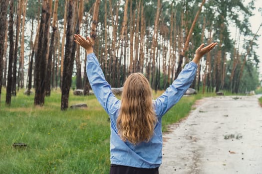 Back view young woman walk in blue raincoat enjoying the woods in park. Open arms outdoors in rainy weather forecast. Tourist rest and feel freedom. Autumn season