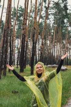 Smiling woman in yellow raincoat walking in autumn forest enjoying rainy weather outdoors. Female tourist discover park in rainy season. Closeness to nature. Mental healing fulfillment clean air