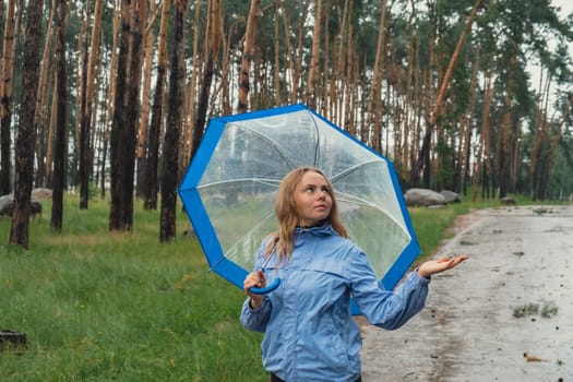 Young woman holding transparent blue umbrella outdoors in forest. Rainy weather day using umbrella. Woman with hand checking how long it will be raining. Forecast