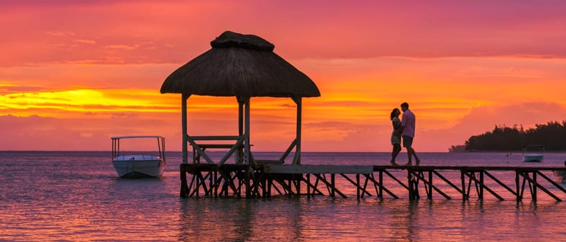Man and Woman watching sunset on a wooden pier in the ocean on a tropical beach in Mauritius, a couple on a honeymoon vacation in Mauritius
