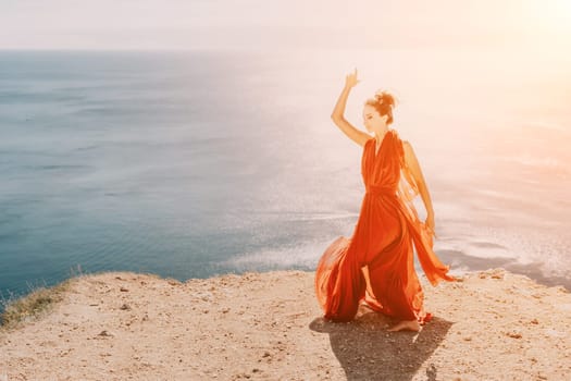 Side view a Young beautiful sensual woman in a red long dress posing on a rock high above the sea during sunrise. Girl on the nature on blue sky background. Fashion photo.