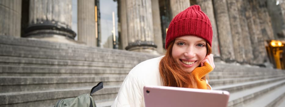 Close up portrait of cute redhead european girl, sitting in red hat near municipal building on stairs, holds digital tablet, reads e-book or surfs internet.