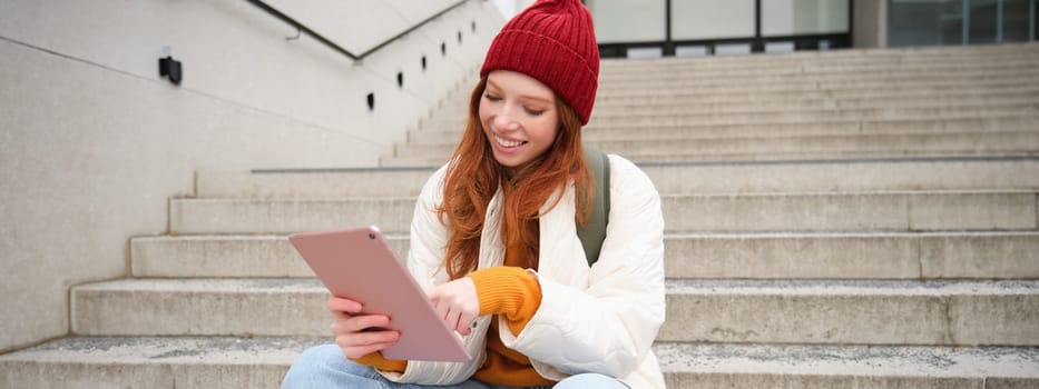 Happy stylish redhead girl, student in red hat, holds digital tablet, uses social media app, searches something online, connects to wifi.