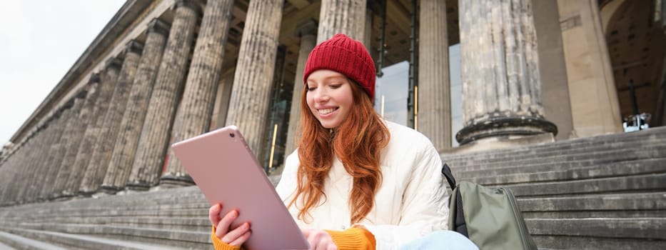 Outdoor shot of young stylish redhead girl sits on staircase and connects to public wifi, uses digital tablet, reads news on gadget.