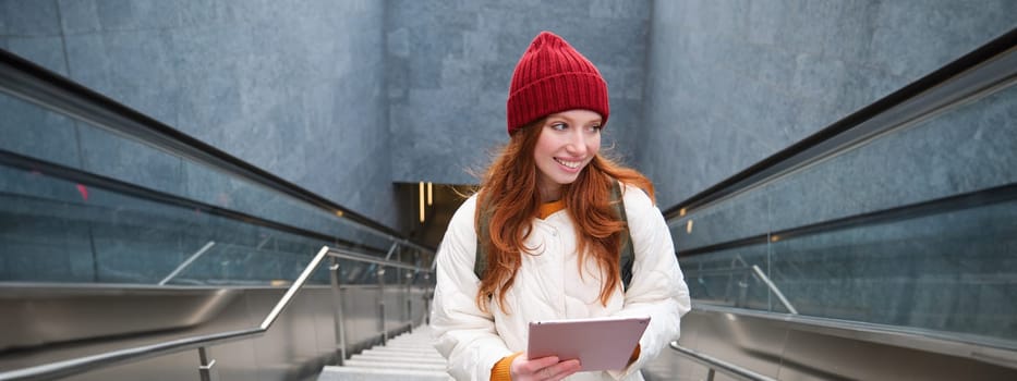 Beautiful redhead female model posing in city, walking up stairs with digital tablet, using gadget to plan her route, reading while going somewere.