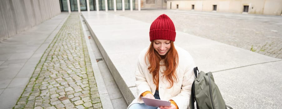 Beautiful redhead woman in red hat, sits with backpack and thermos, using digital tablet outdoors, connects to wifi, texts message, books tickets online.