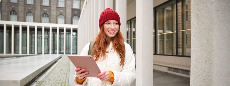 Stylish ginger girl, tourist walks with digital tablet around city, woman connects to iternet on her gadget, looking up information, texting message.