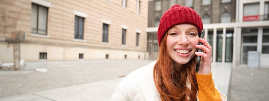 Stylish modern redhead girl talks on mobile phone, makes a telephone call, calling someone on smartphone app from outside, stands on street and smiles.