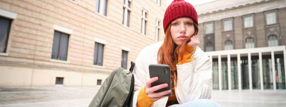 Redhead 20s years girl, sits with smartphone outside building, looks complicated and upset, frowns thoughtful, holds mobile phone in hand.