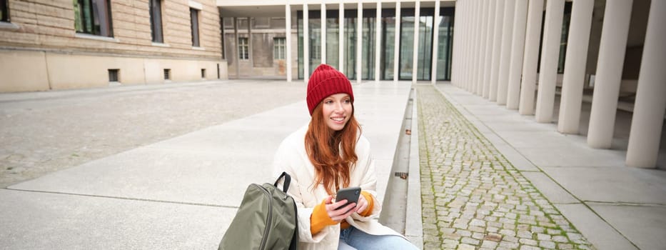 Social media and people. Young redhead girl sits on street, uses mobile phone app, looks up information in internet, holds smartphone.