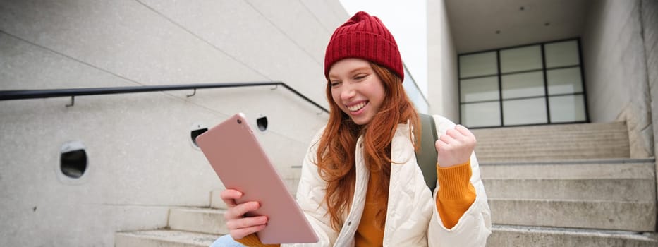 Young stylish girl, redhead female students sits on stairs outdoors with digital tablet, reads, uses social media app on gadget, plays games while waits on street.