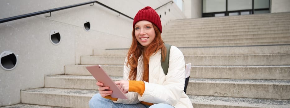 Young stylish girl, redhead female students sits on stairs outdoors with digital tablet, reads, uses social media app on gadget, plays games while waits on street.