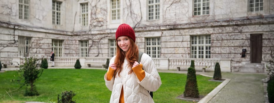 Portrait of smiling girl, backpacker in coat and red hat, wearing warm clothes for tourist trip around Europe in winter, walking around historical building.