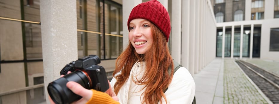 Smiling redhead girl photographer, taking pictures in city, makes photos outdoors on professional camera. Young talent and hobby concept