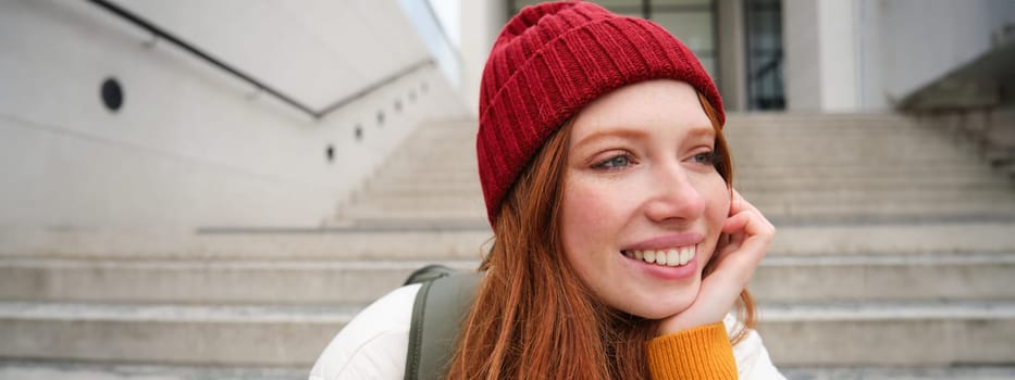 Close up portrait of beautiful redhead girl in red hat, urban woman with freckles and ginger hair, sits on stairs on street, smiles and looks gorgeous.