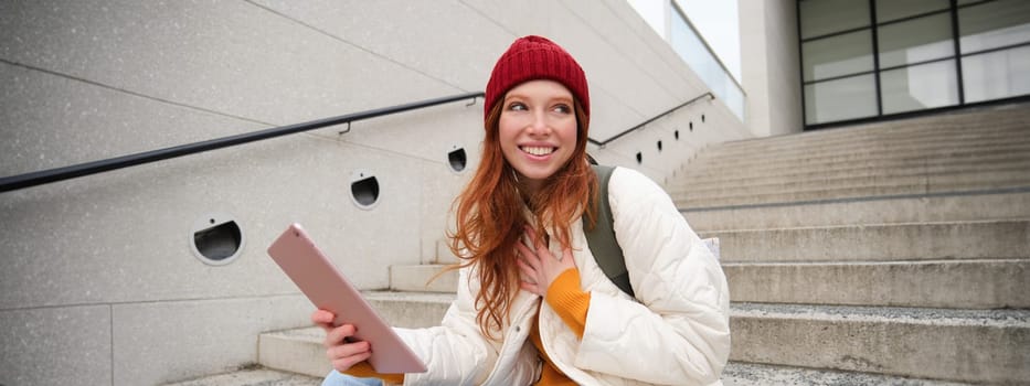 Young stylish girl, redhead female students sits on stairs outdoors with digital tablet, reads, uses social media app on gadget, plays games while waits on street.