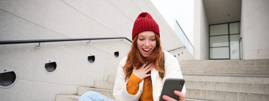 Image of redhead girl with surprised face expression, looks amazed at her smartphone, reads great news on phone, sits on street stairs.