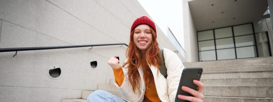 Joyful young redhead girl sits on street stairs, say yes, wins and celebrates with phone in hand, receive positive news on her mobile application and rejoice.