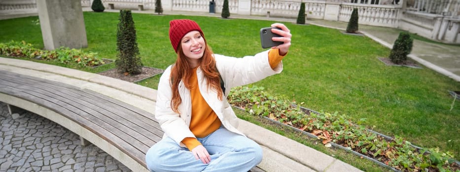 Young teen redhead girl sits on bench in park and takes selfie, makes a photo of herself with smartphone app, records vlog.