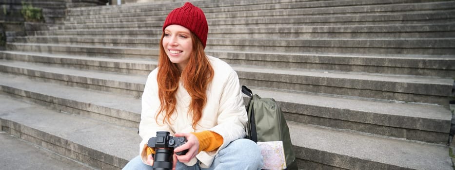 Young student, photographer sits on street stairs and checks her shots on professional camera, taking photos outdoors.