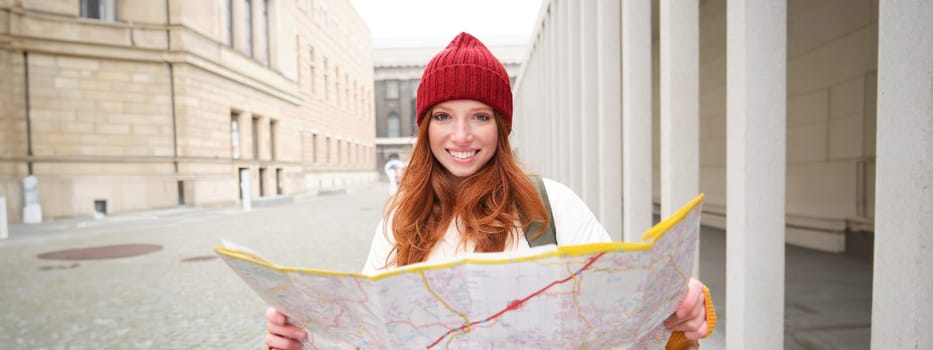 Beautiful redhead woman, tourist with city map, explores sightseeing historical landmark, walking around old town, smiling happily.