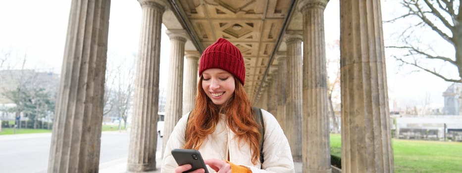 Mobile broadband and people. Smiling redhead 20s girl with backpack, uses smartphone on street, holds mobile phone and looks at application.
