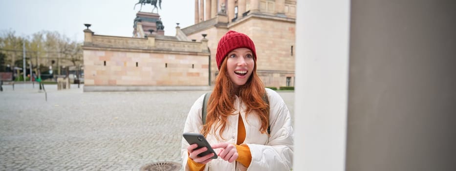 Tourism and sightseeing concept. Young redhead woman, tourist walks around city, looks at her smartphone app and at history stand, explores adventures.