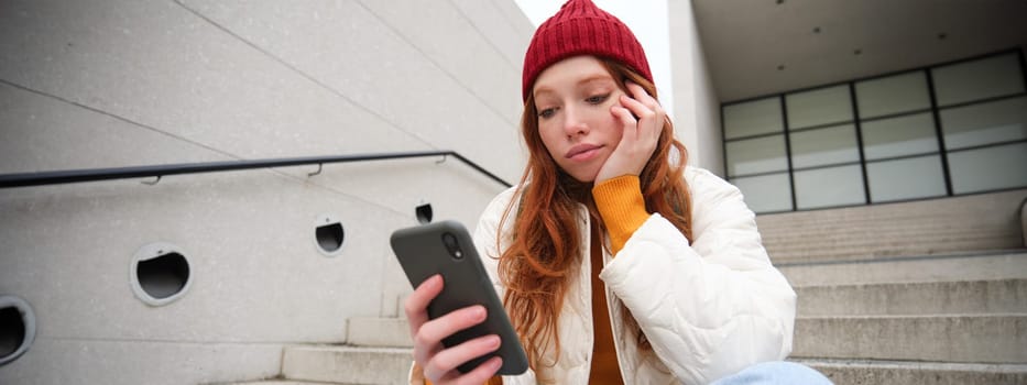 Mobile phones and people. Young stylish redhead girl sits on stairs with telephone, uses smartphone app, reads smth online.