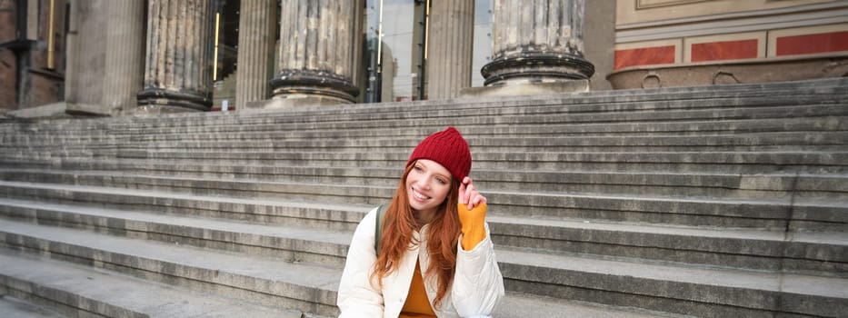 Stylish young redhead woman, talking on mobile phone app, using social media application, looking for something online on smartphone, sits on stairs outdoors.