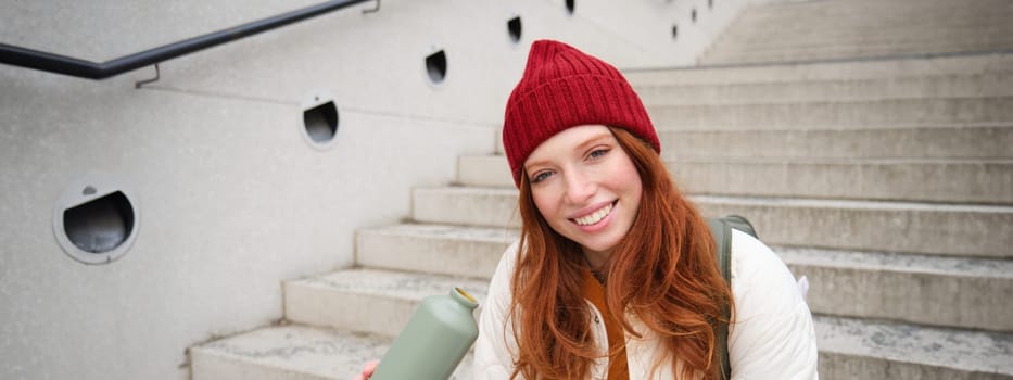 Happy redhead woman, tourist with thermos, drinks her hot tea, coffee from travel flask, restests during her travelling in city and sightseeing.