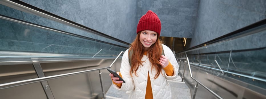 Outdoor shot of young woman plans route, follows map on smartphone app, goes up stairs with backpack and smiles.
