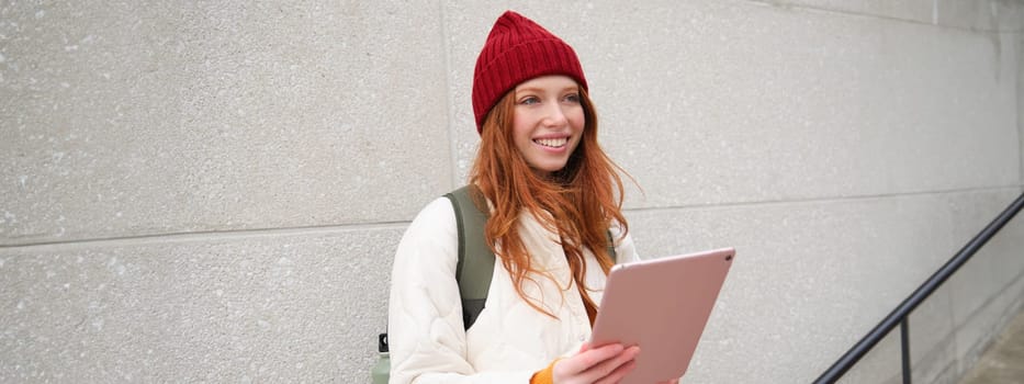 Young redhead girl, walks with digital tablet around city, stands on street with backpack and gadget, looks at online map, searches for a place in internet.