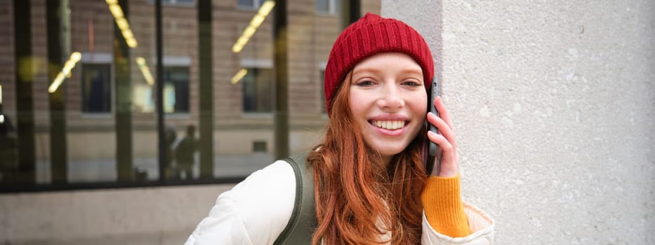 Young people and mobile connection. Happy redhead girl talks on phone, makes telephone call, stands outdoors with backpack and uses smartphone app.