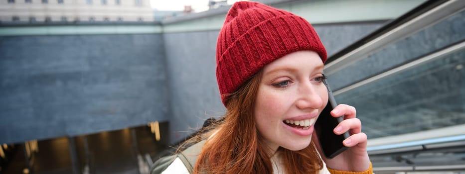 Portrait of young redhead woman walks around city, goes up stairs with mobile phone, talks on smartphone and smiles.