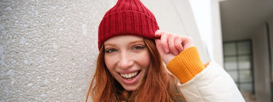 Stylish redhead girl in red hat, smiles and looks happy, poses outdoors on street, looks relaxed and lively.