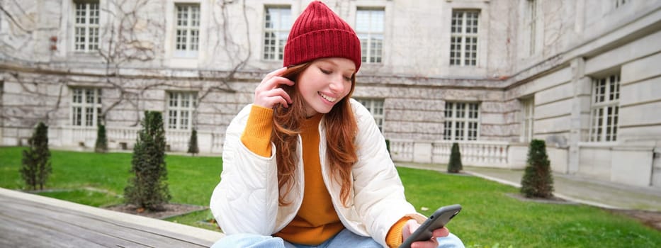 Young smiling redhead girl sits on bench and uses smartphone app, reads news online, watches video on mobile phone while relaxed in park.