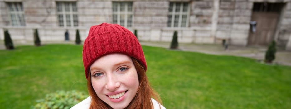 Vertical portrait of cute redhead female student in red hat and warm gloves, sits in park on bench, smiles and looks cute at camera.