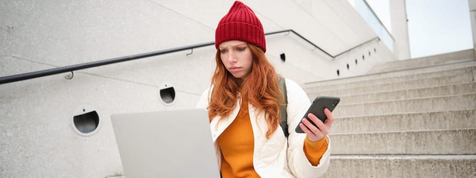 Portrait of redhead woman sits on stairs, uses laptop and holds smartphone, looks confused and upset at computer screen, tries to connect to wifi public internet.