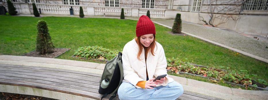 Young redhead girl student, sits on bench and uses smartphone app, watches videos online, sends message on mobile phone.