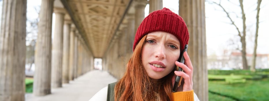 Portrait of redhead girl in red hat, calls someone, listens to voice message with concerned, confused face expression, using smartphone.