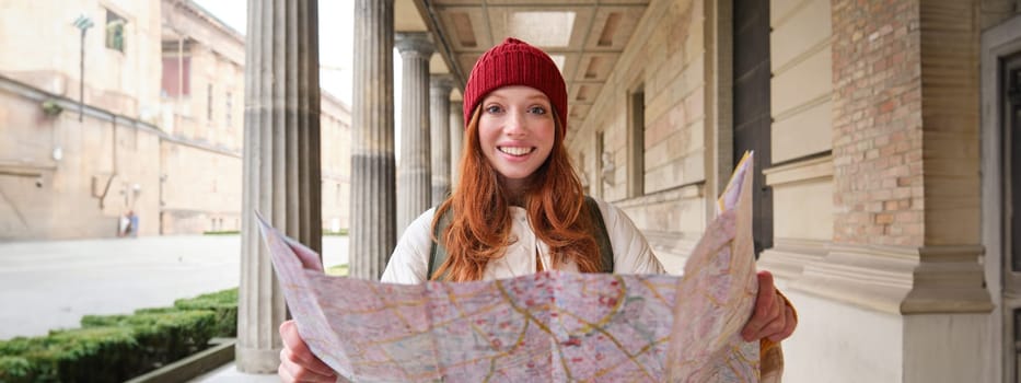 Smiling young redhead woman in red hat, looks at paper map to look for tourist attraction. Tourism and people concept. Girl explores city, tried to find way.