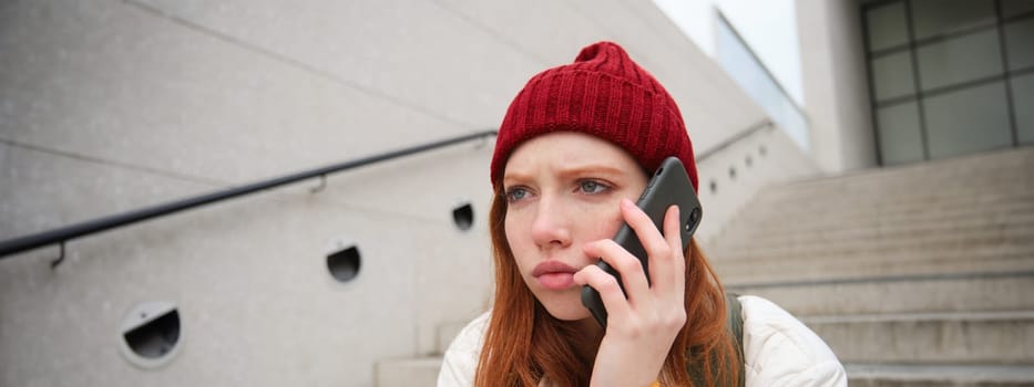Redhead girl with concerned face, looking worried while answering phone call, hears bad news over telephone conversation, rings someone with upset emotion.