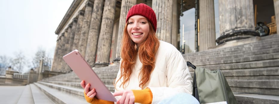 Outdoor shot of young stylish redhead girl sits on staircase and connects to public wifi, uses digital tablet, reads news on gadget.