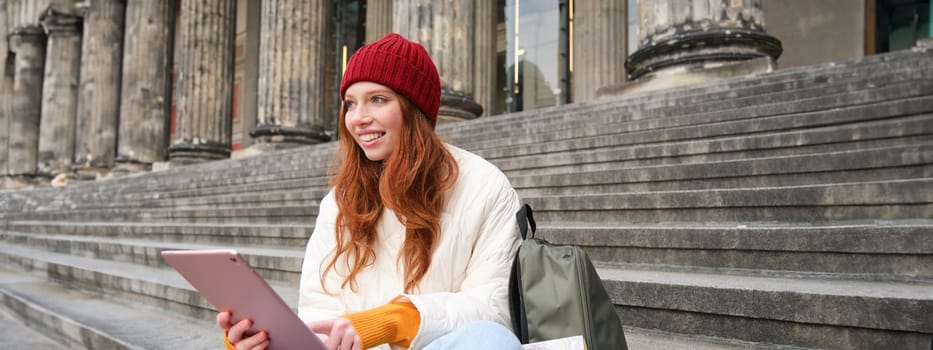 Beautiful young modern girl with red hair, holds digital tablet, sits on stairs near museum and connects public internet, sends message on gadget app.