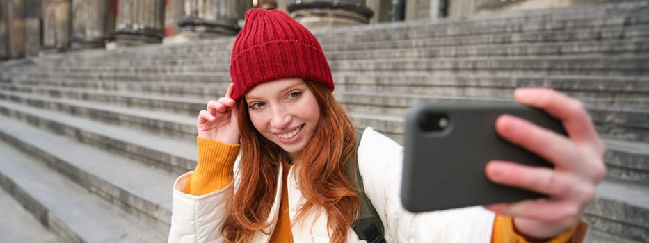 Stylish young girl in red hat, takes photos on smartphone camera, makes selfie as she sits on stairs near museum, posing for photo with app filter.