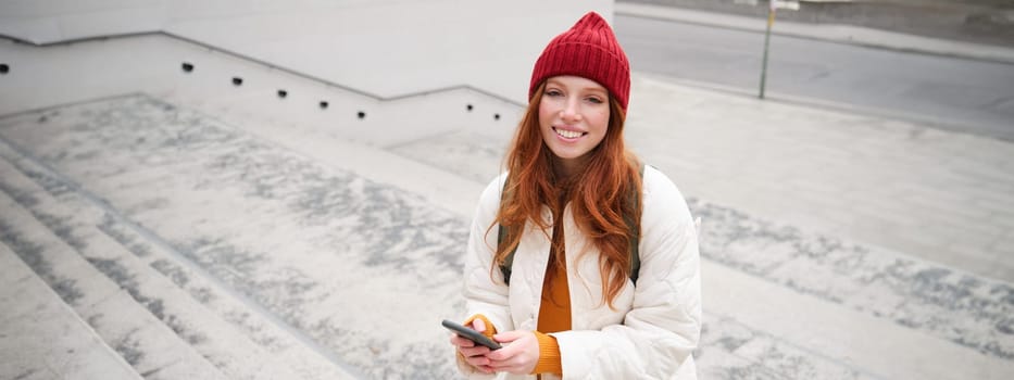 Smiling redhead girl, student tourist walks around city, goes up the stairs, looks at mobile phone map to get around town, sends message on smartphone.