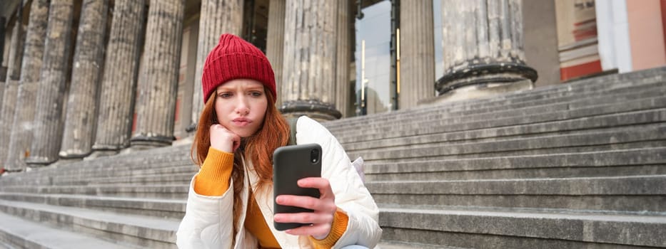 Portrait of young redhead woman with complicated face, sits on street stairs in red hat, holds smartphone and frowns thoughtful, feels uneasy.